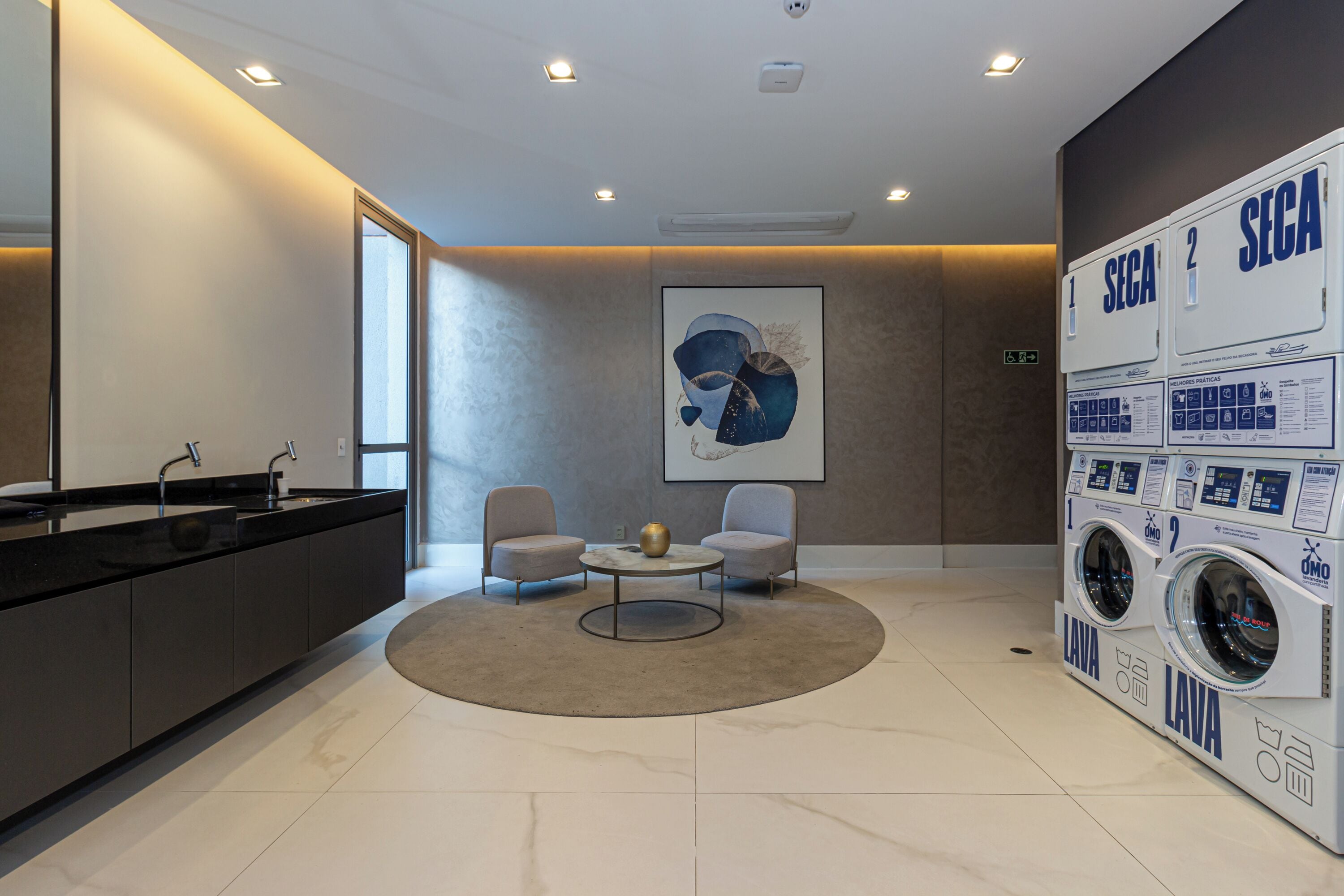 A modern hotel lobby with a black reception desk, two armchairs, a coffee table, a large abstract painting, and a wall-mounted laundry machine.