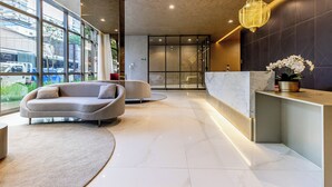 A modern reception area with a marble reception desk, a sofa, and a large window offering a view of the street. - Charlie Grand Vitrali Moema (São Paulo)