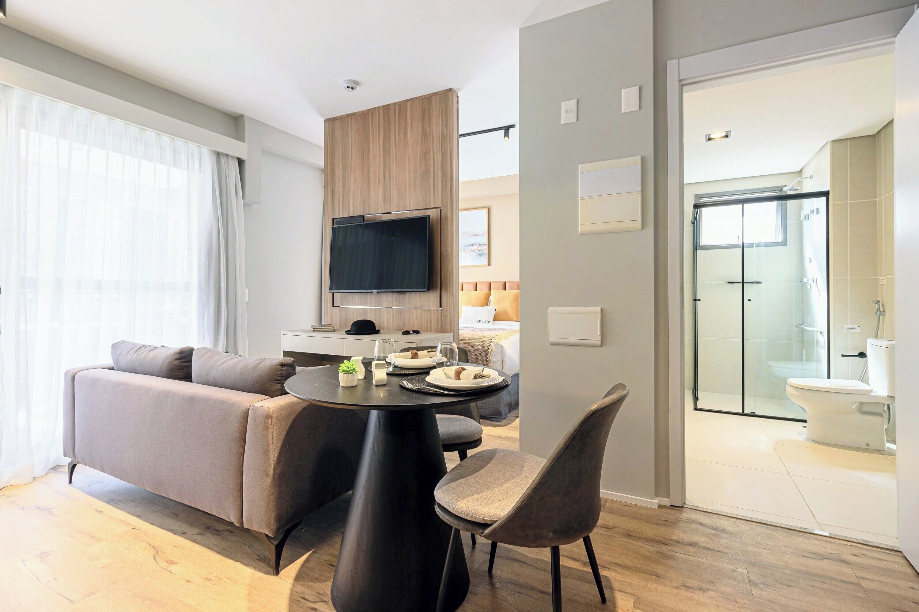 A modern living room with a sofa, dining table, and chairs, a TV mounted on a wooden panel, and a glass shower door leading to a bathroom.