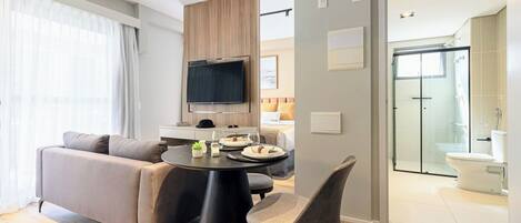 A modern living room with a sofa, dining table, and chairs, a TV mounted on a wooden panel, and a glass shower door leading to a bathroom.
