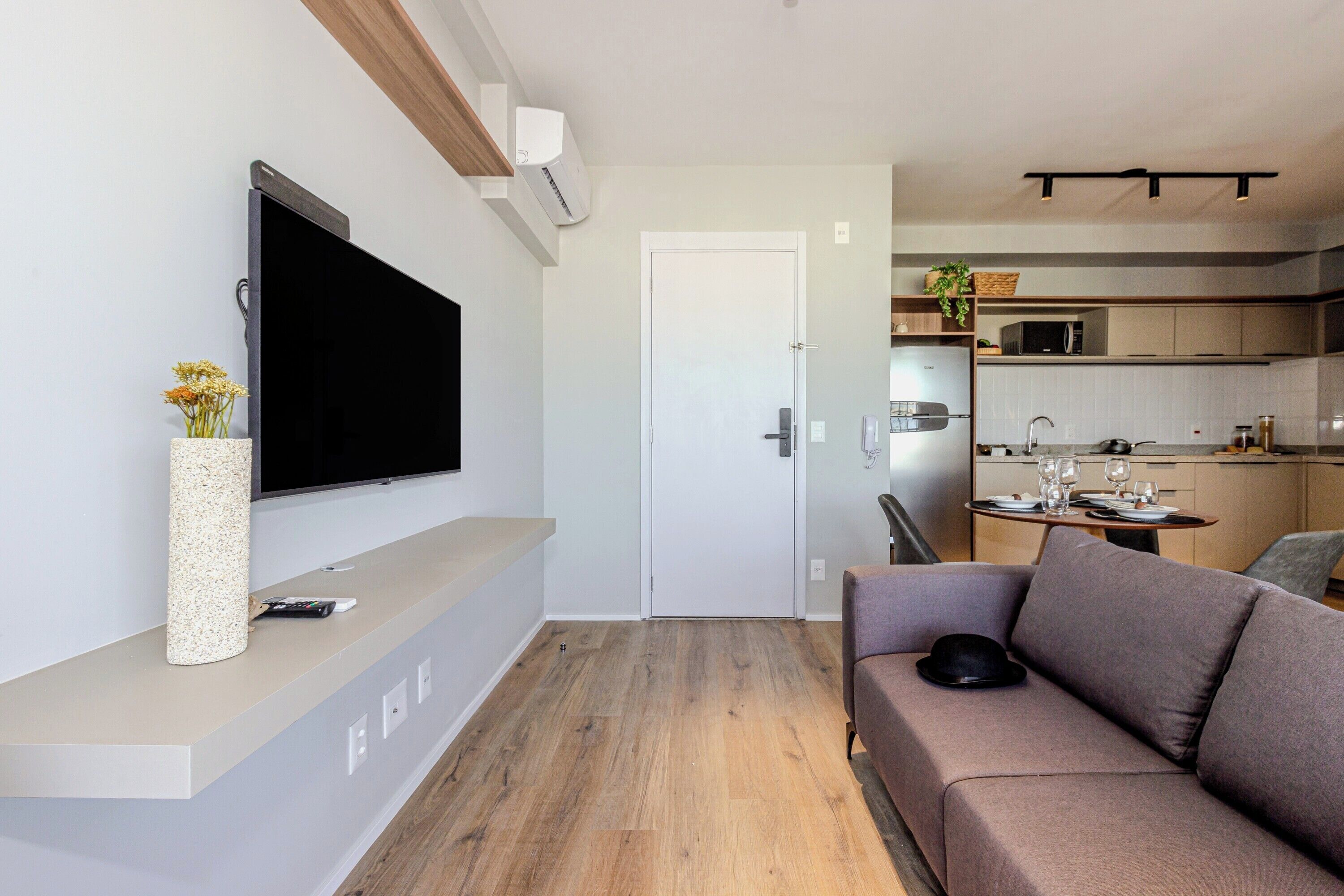 A modern living room with a flat-screen TV, a sofa, and a coffee table. There is a kitchen area with a microwave and a sink visible through an open door.