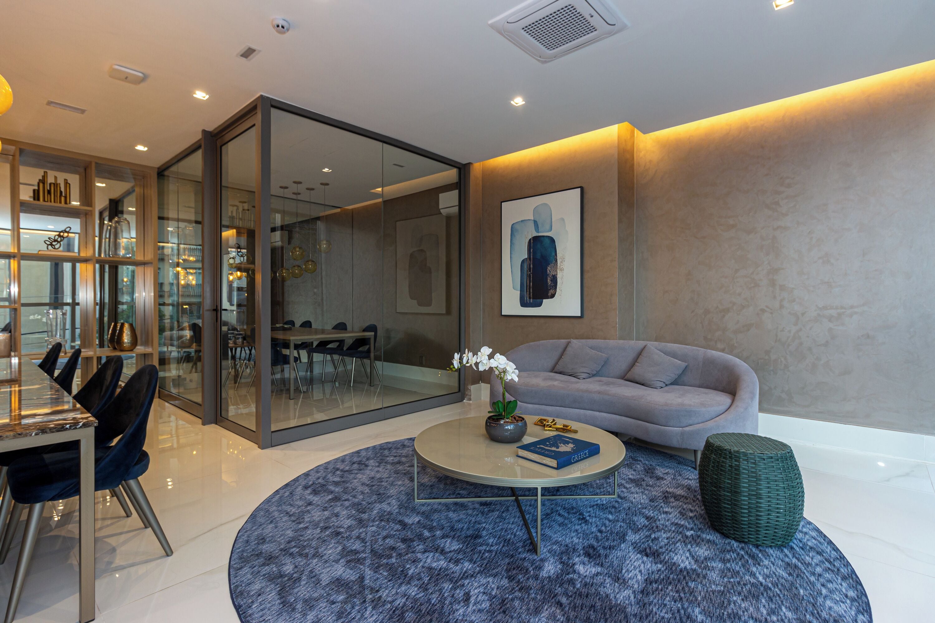 A modern living room with a round coffee table, a grey sofa, and a glass partition separating the living area from a dining space.