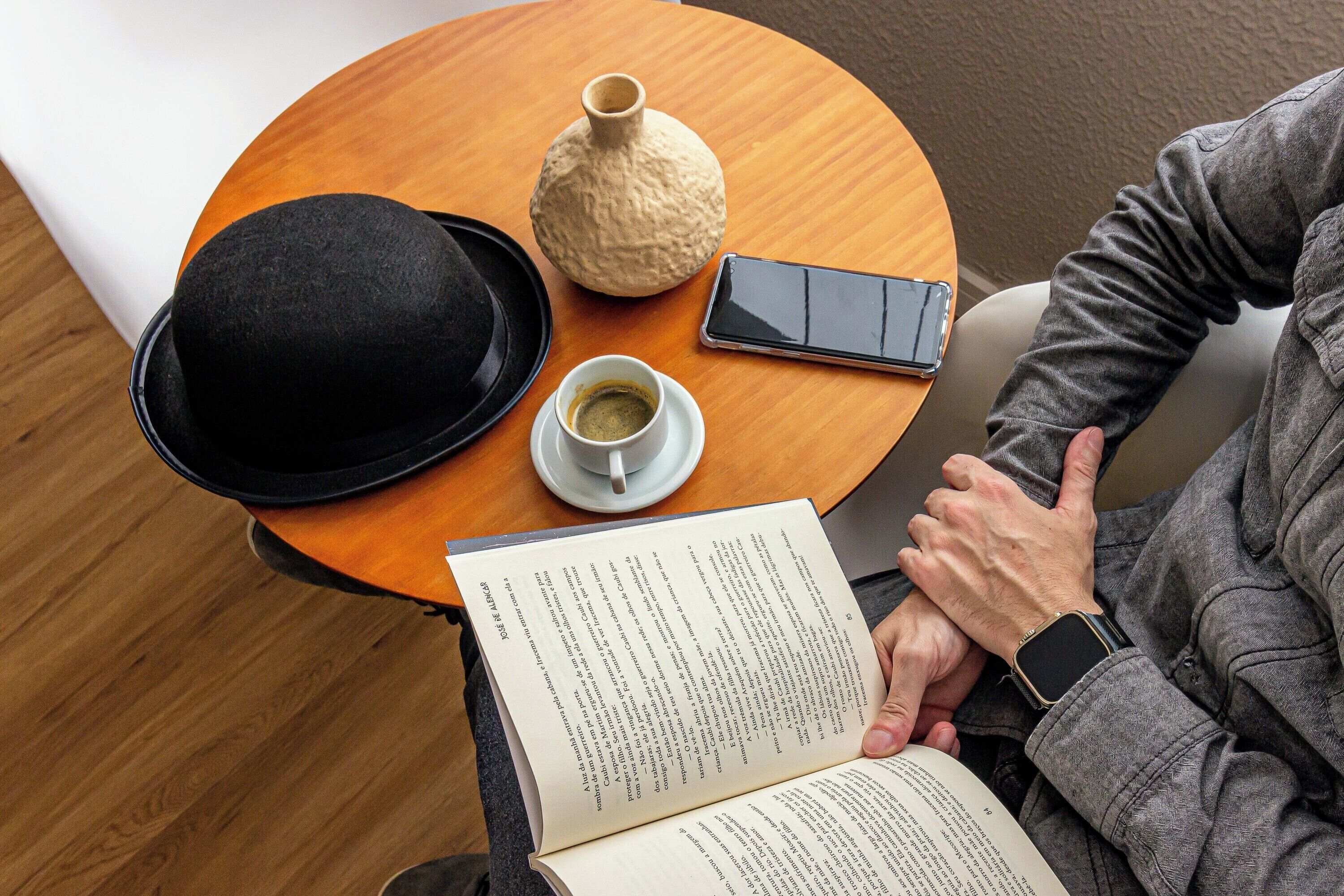 Iemand leest een boek aan een ronde houten tafel, met een zwarte hoed, een witte koffiekop en een smartphone.