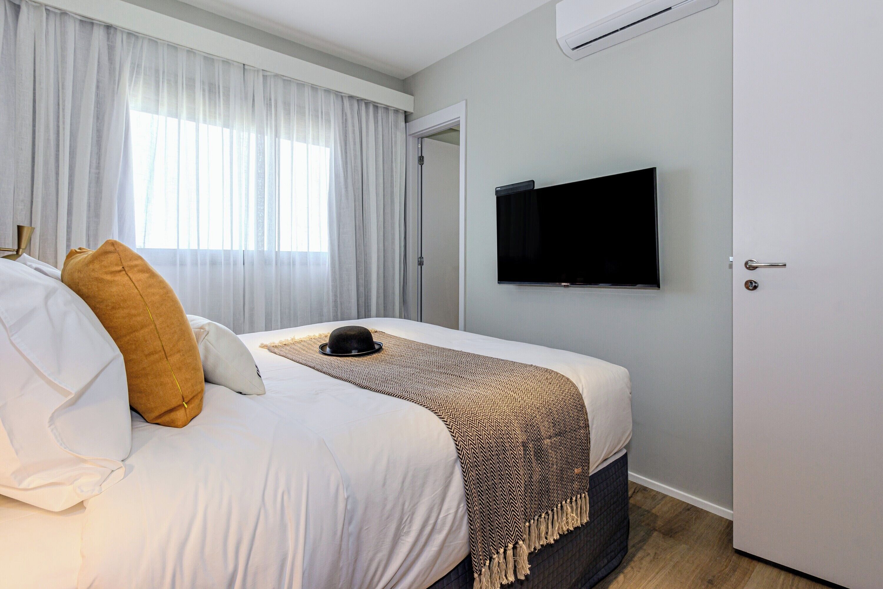 Een netjes opgemaakt bed met wit en bruin beddengoed, een aan de muur bevestigde televisie en een deur aan de rechterkant.