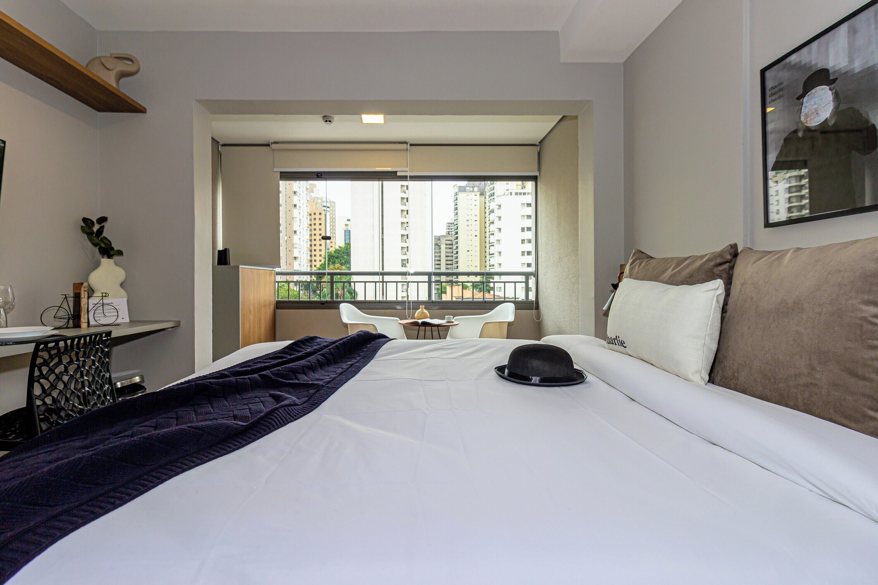 Habitación de hotel con una cama grande, un balcón con vistas a edificios altos, un escritorio con una silla y una mesita con un florero y una bicicleta.