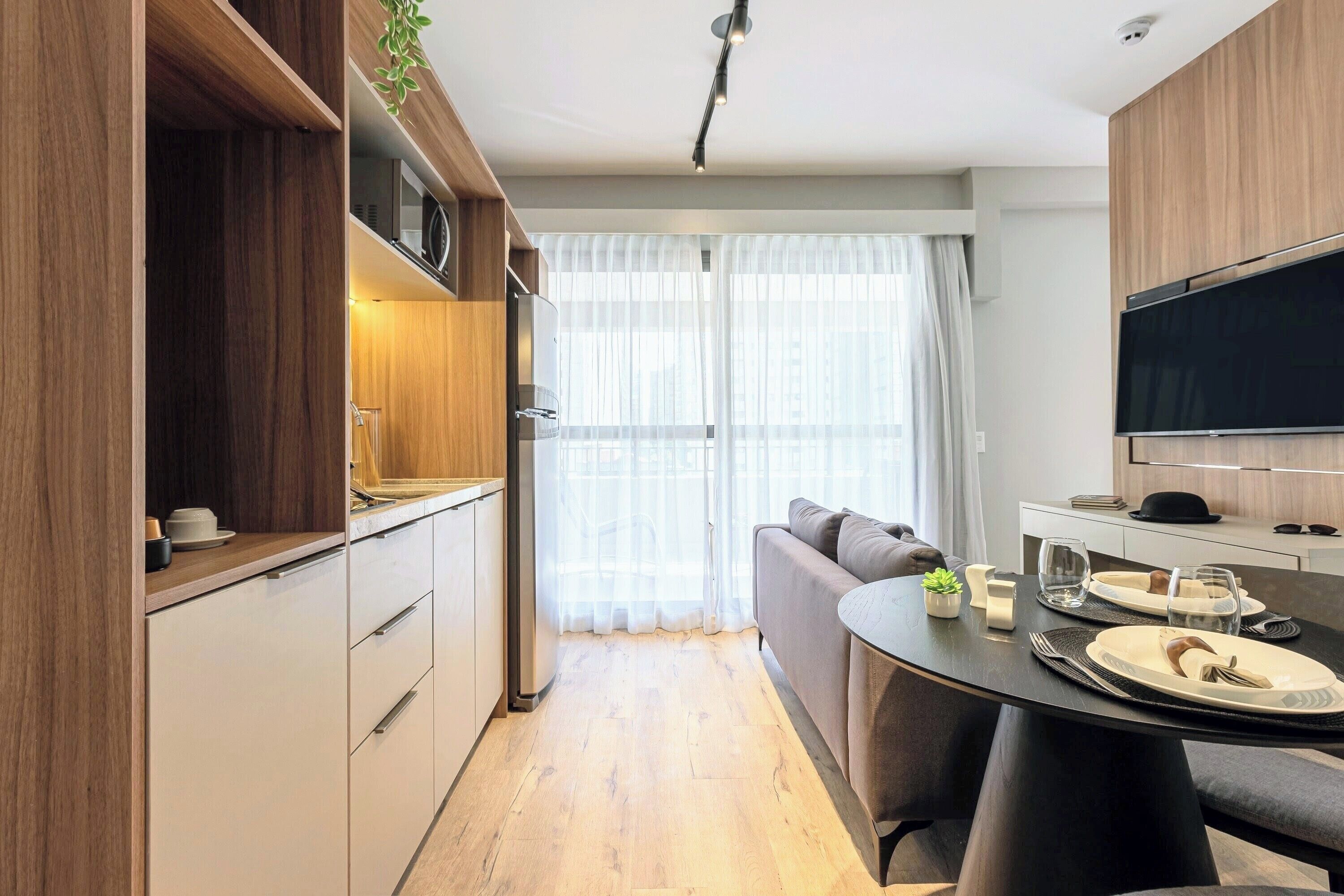A modern kitchen with wooden cabinets, a dining table set for two, and a large TV mounted on the wall.