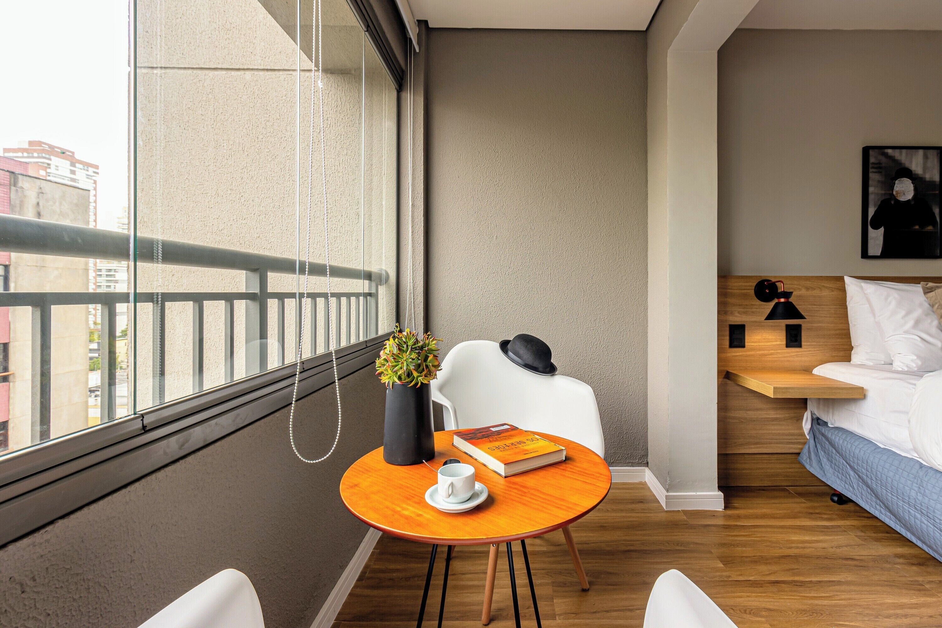 A modern hotel room with a wooden floor, a round table with a white cup and a book, a white chair, a bed with a wooden headboard, and a city view through a large window.