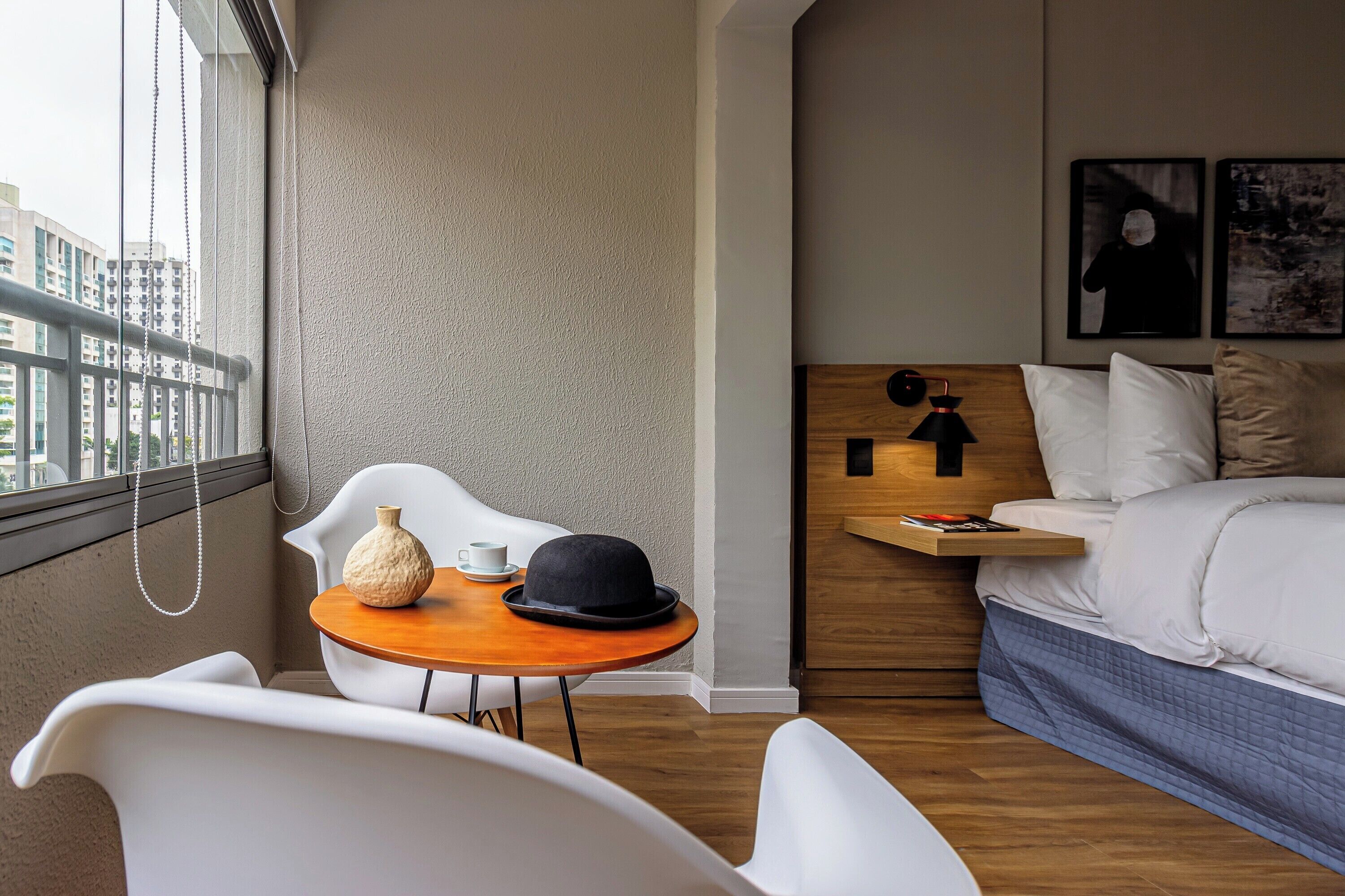 A hotel room with a bed, a bedside table with a lamp, a small table with a vase and a hat, a chair, and a view of the city through the window.