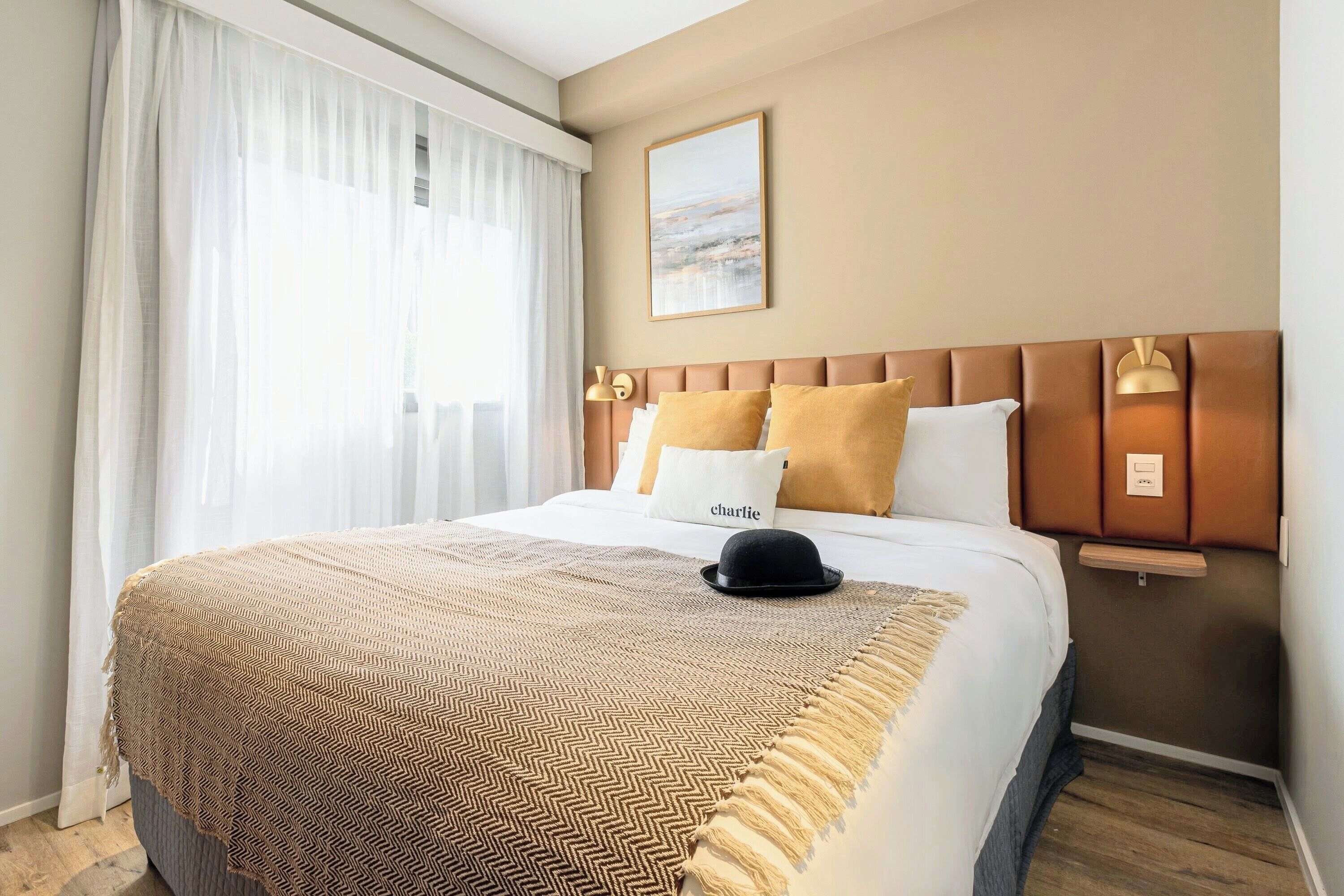 Habitación de hotel con cama, cabecero, mesita de noche, lámpara y ventana con cortinas.