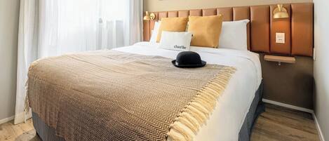 Habitación de hotel con cama, cabecero, mesita de noche, lámpara y ventana con cortinas.