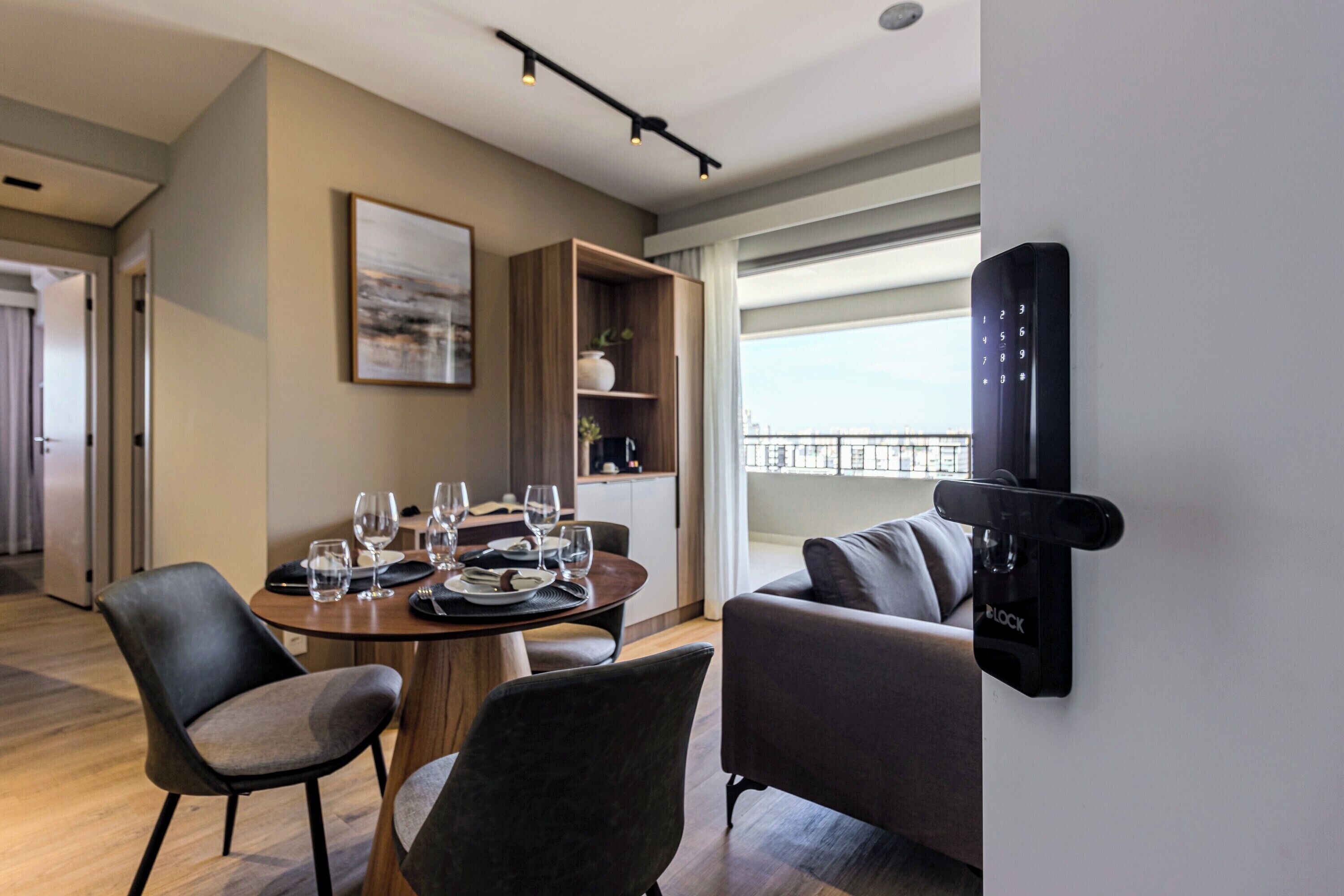 Un comedor moderno con una mesa redonda dispuesta para cuatro personas, rodeado de sofás oscuros. Se observa una estantería y un cuadro en la pared.