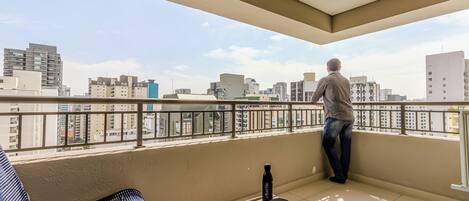 A balcony with a view of the cityscape, a chair, a table with a bottle and a hat, and a person standing by the railing.