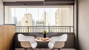 A balcony with two white chairs, a small table with a book and a plant, and a city view through large windows. - Charlie Grand Vitrali Moema (São Paulo)