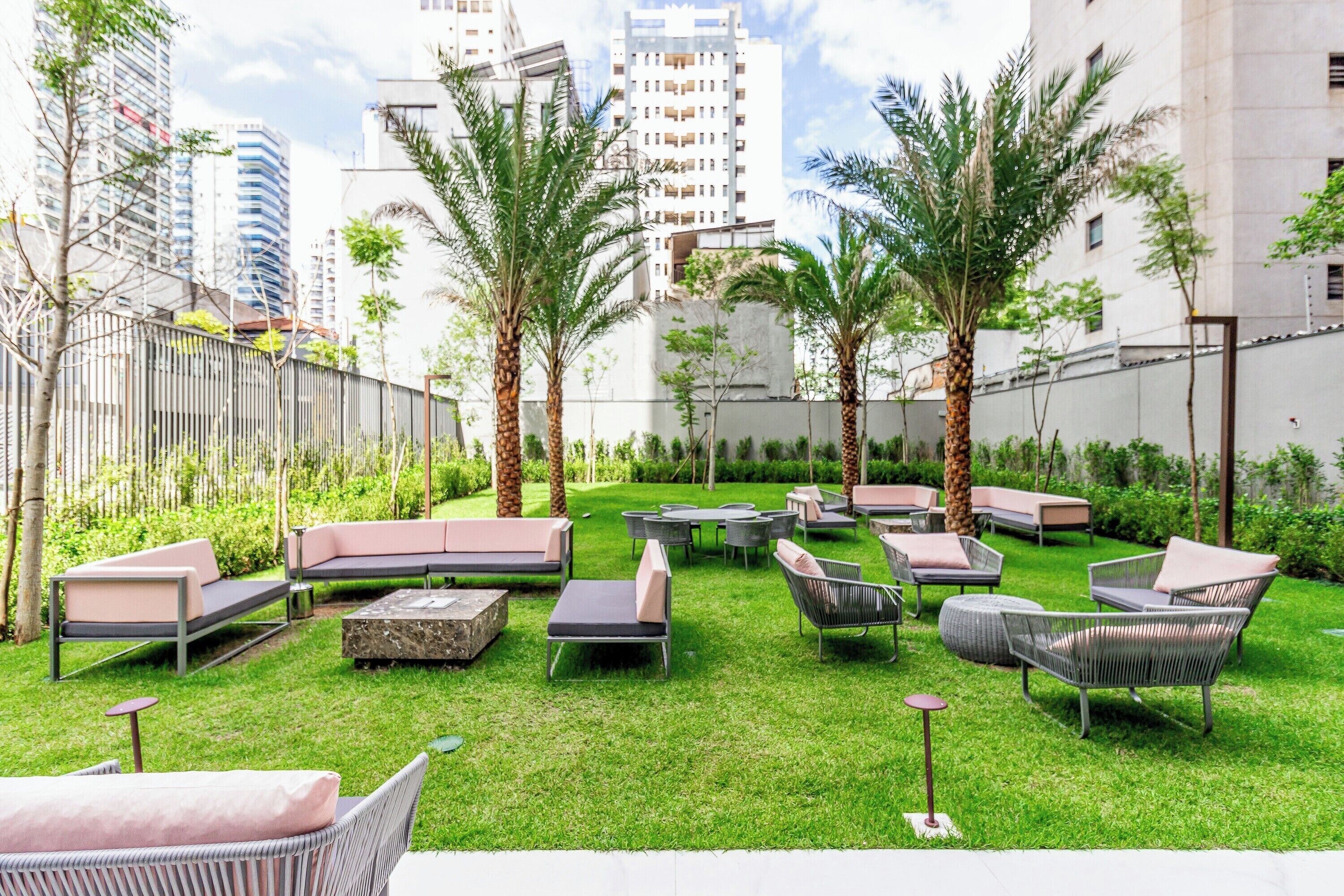 Een buitenverblijf met meerdere lounge fauteuils en tafels, omgeven door palmbomen en groen, met hoge gebouwen op de achtergrond.