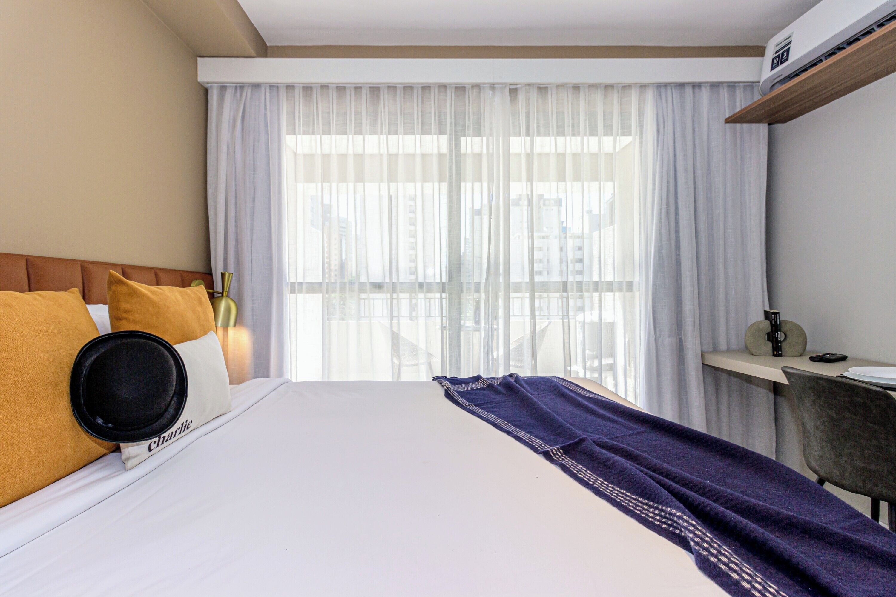 A hotel room with a bed, a window with sheer curtains, a bedside table, and a chair.