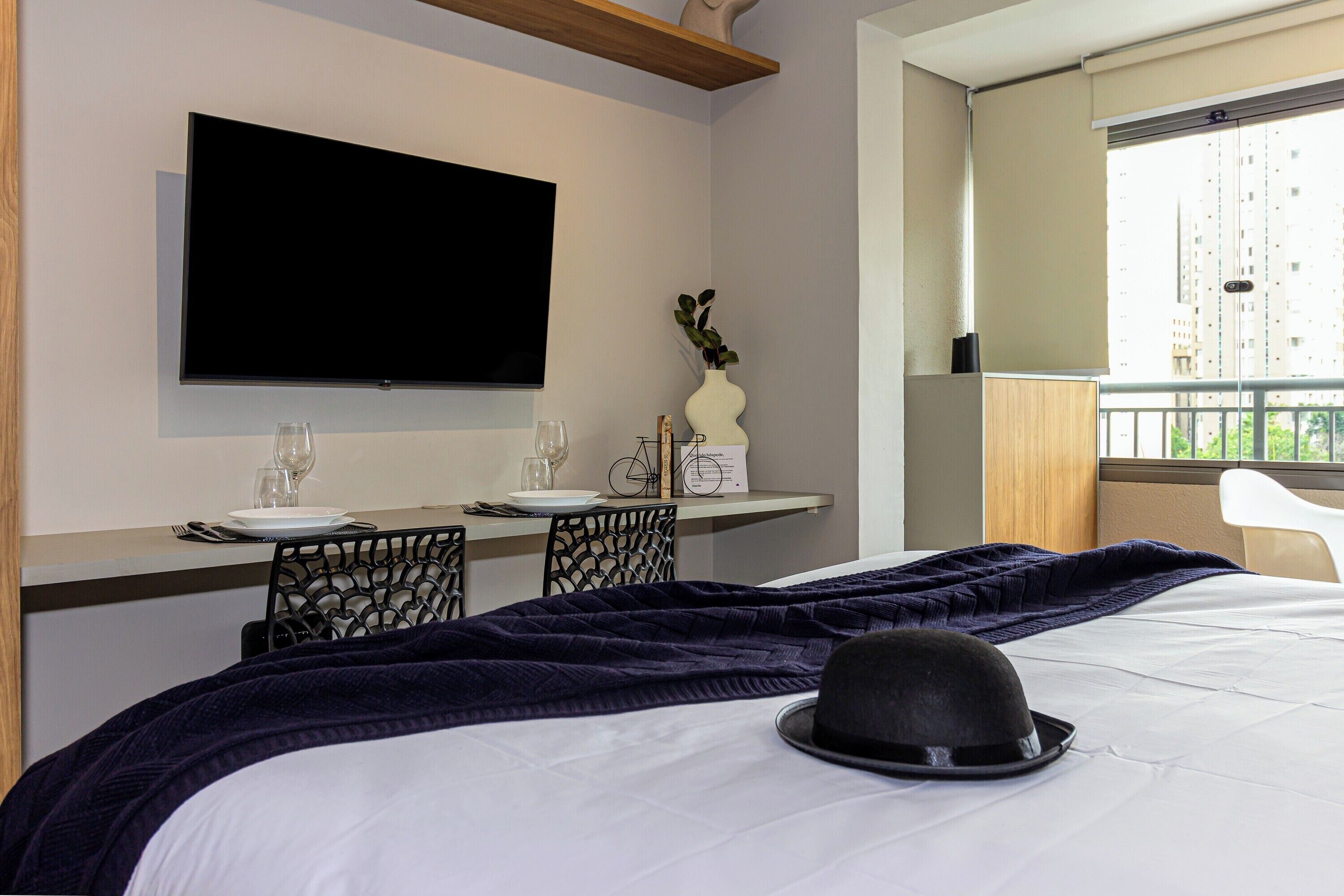Ein Hotelzimmer mit Bett, Wandfernseher, Esstisch mit Stühlen und Fenster mit Blick auf Gebäude.