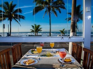 Une table dressée avec de la nourriture et des boissons, donnant sur une plage bordée de palmiers et sur l’océan.