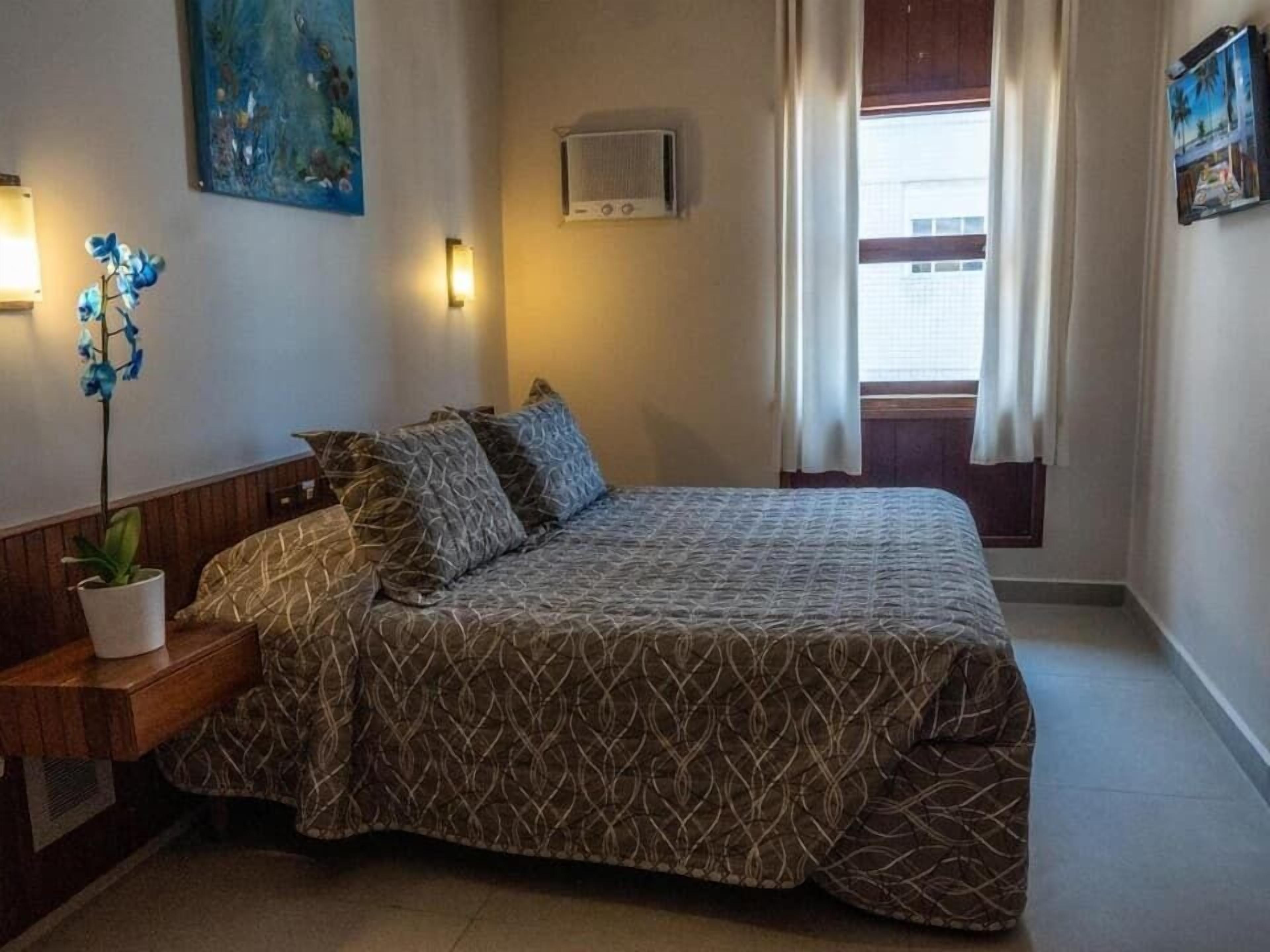 A bedroom with a bed, a nightstand, a potted plant, a wall-mounted air conditioner, and a window with curtains.