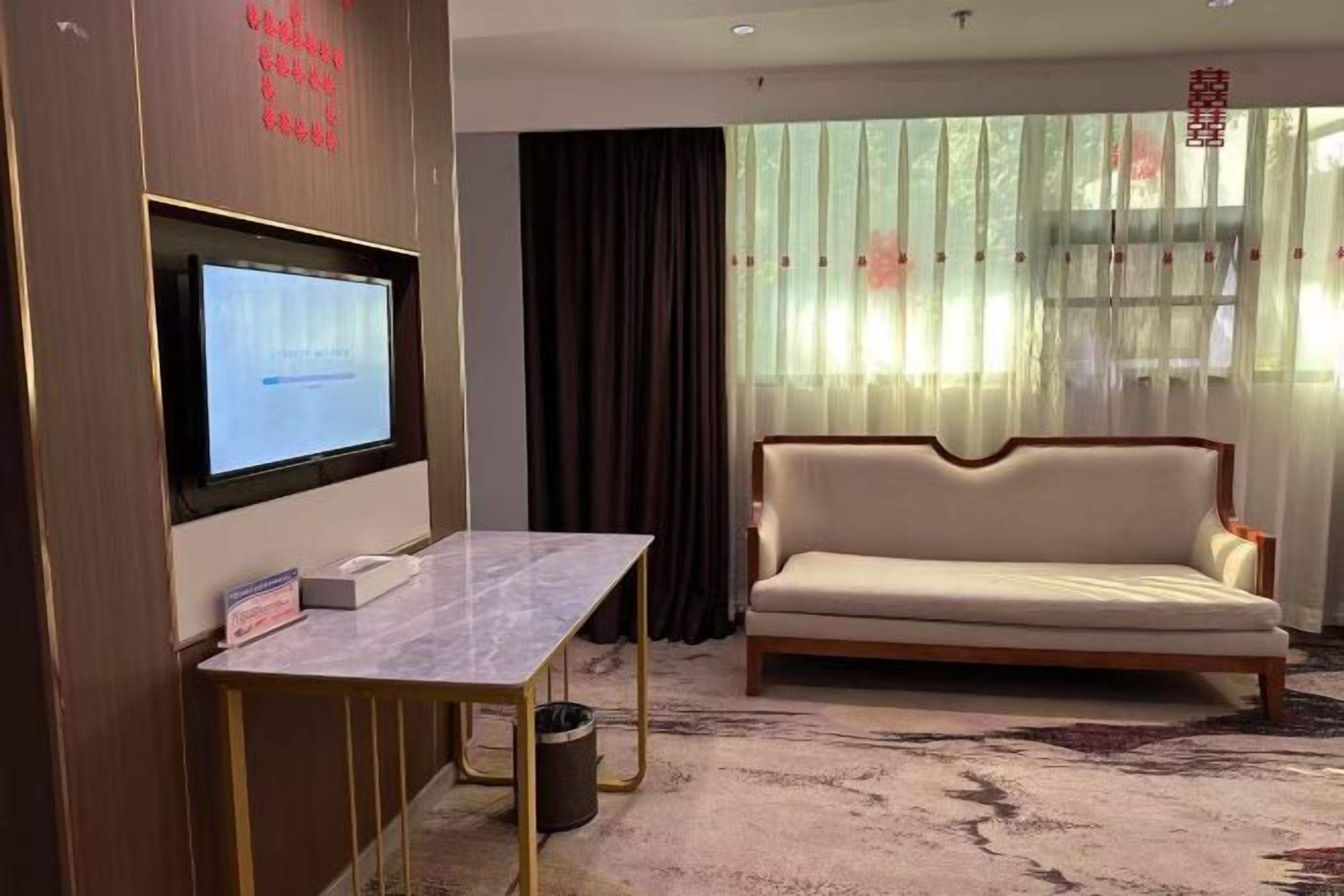 A room with a sofa, a TV mounted on a wall, a small table, and a window with sheer curtains.