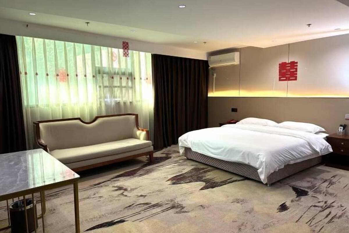 A hotel room with a bed, a sofa, a small table, and a large window with curtains.