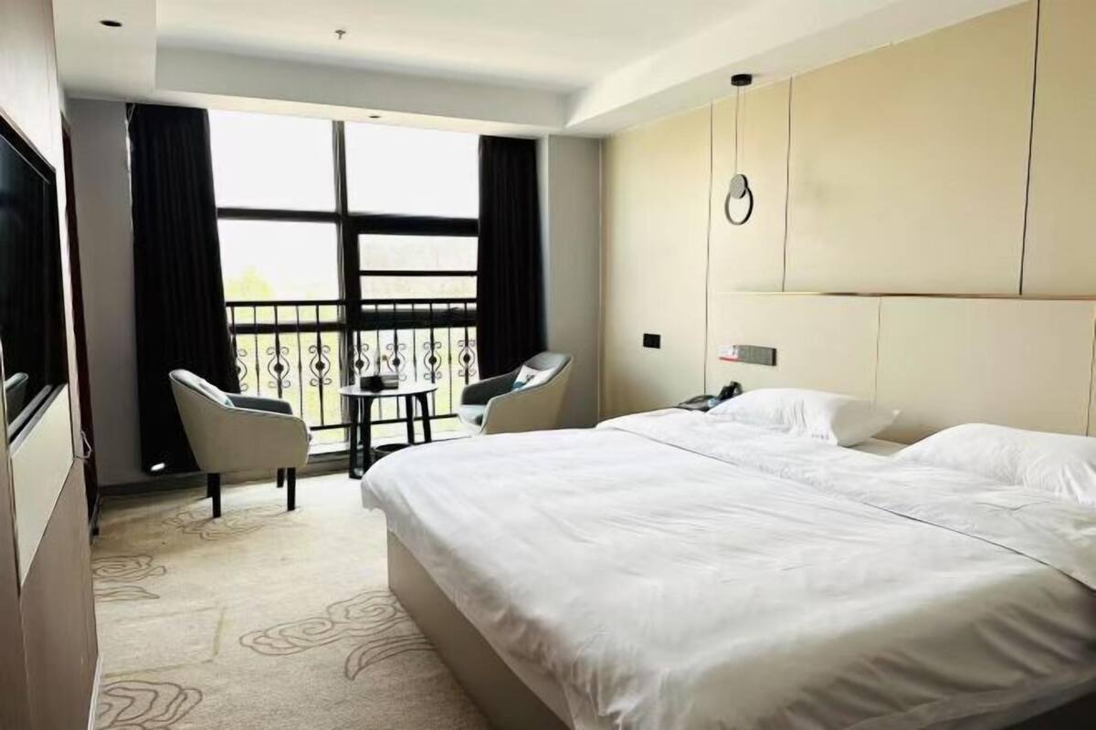 A hotel room with a large bed, two chairs, a small table, a balcony with a railing, and a clock on the wall.