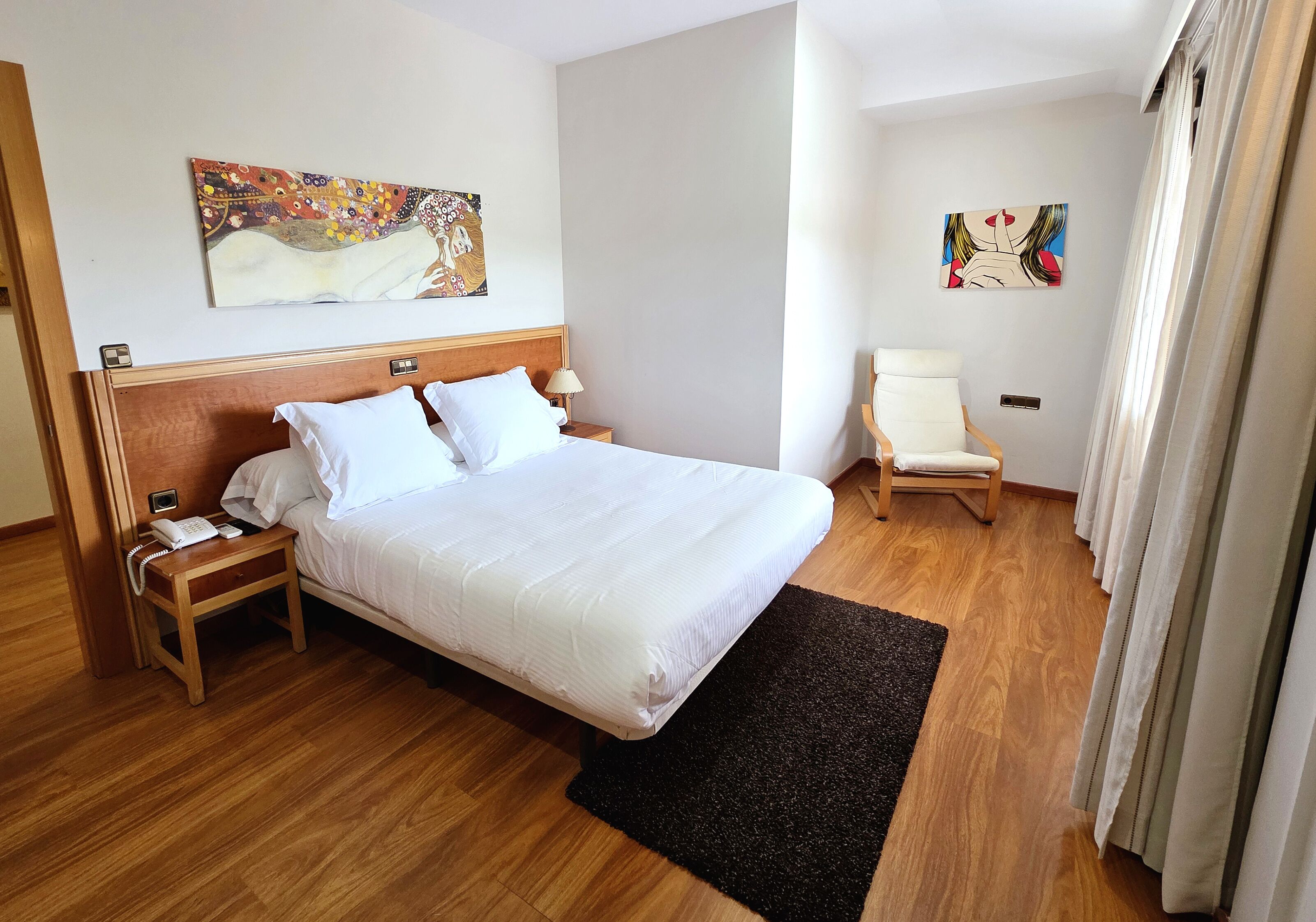 A hotel room with a large bed, a bedside table with a phone, a small chair, and two paintings on the walls.