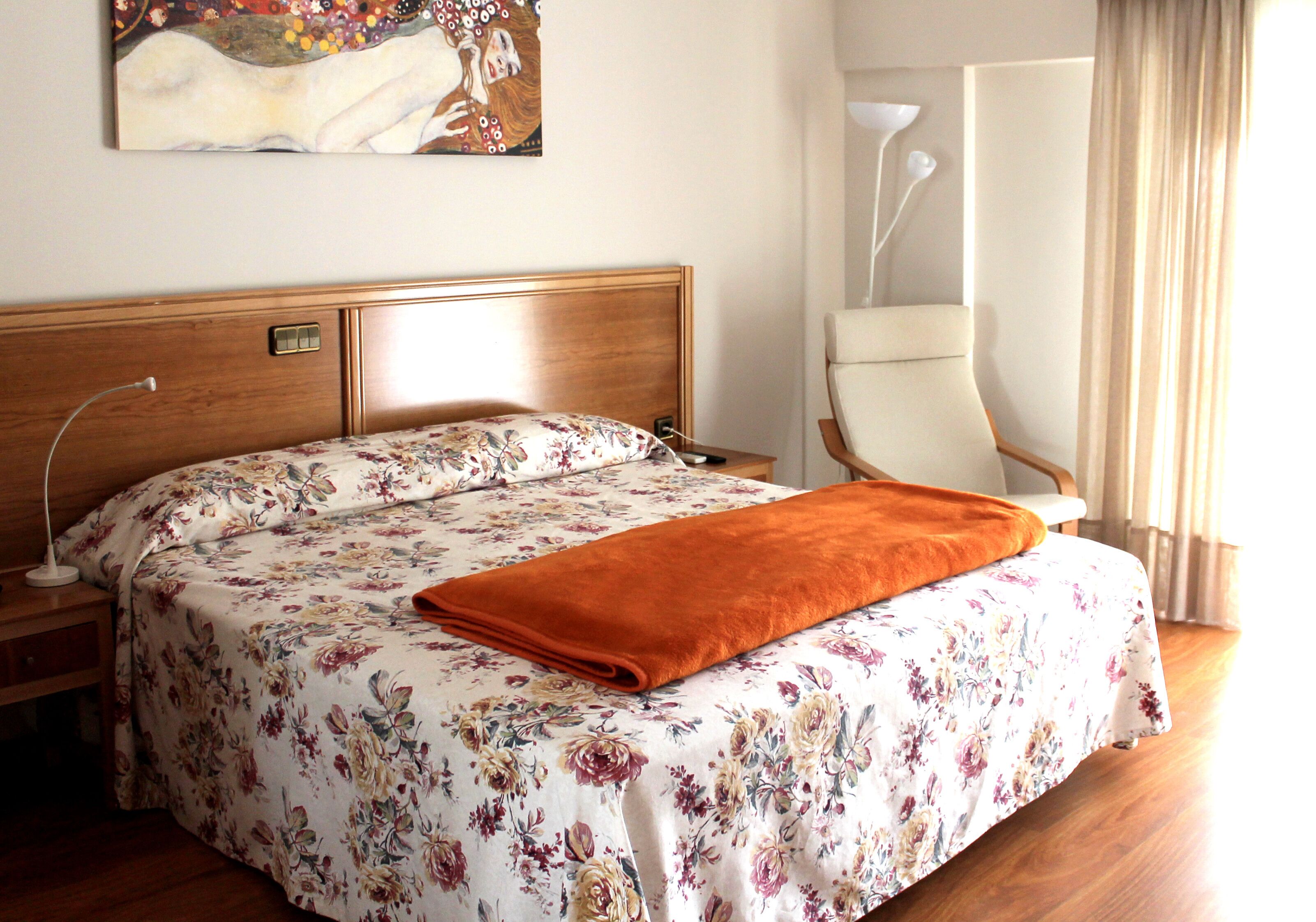 Una cama bien hecha con una colcha floral y una manta naranja, un cabecero de madera, una lámpara de noche y un cuadro en la pared.