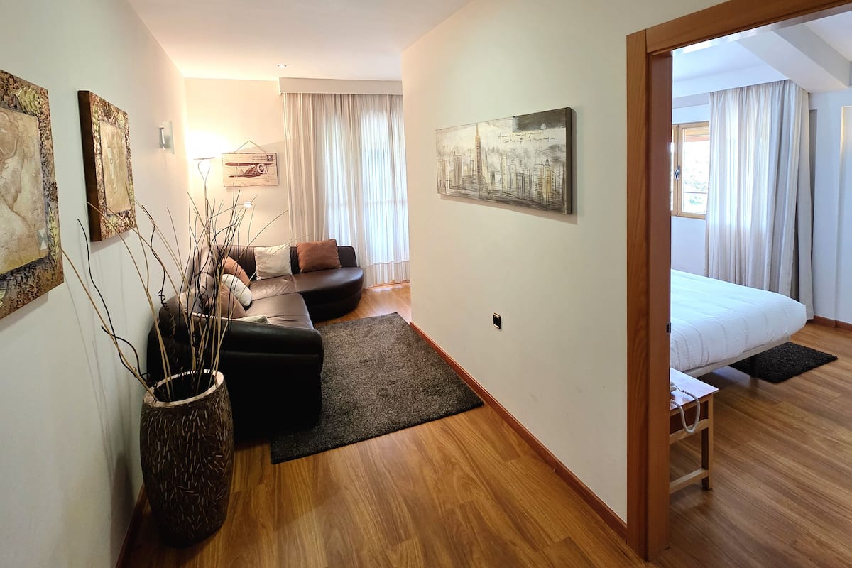 A modern hotel room with a wooden floor, a sofa, a bed, and a large window with curtains.