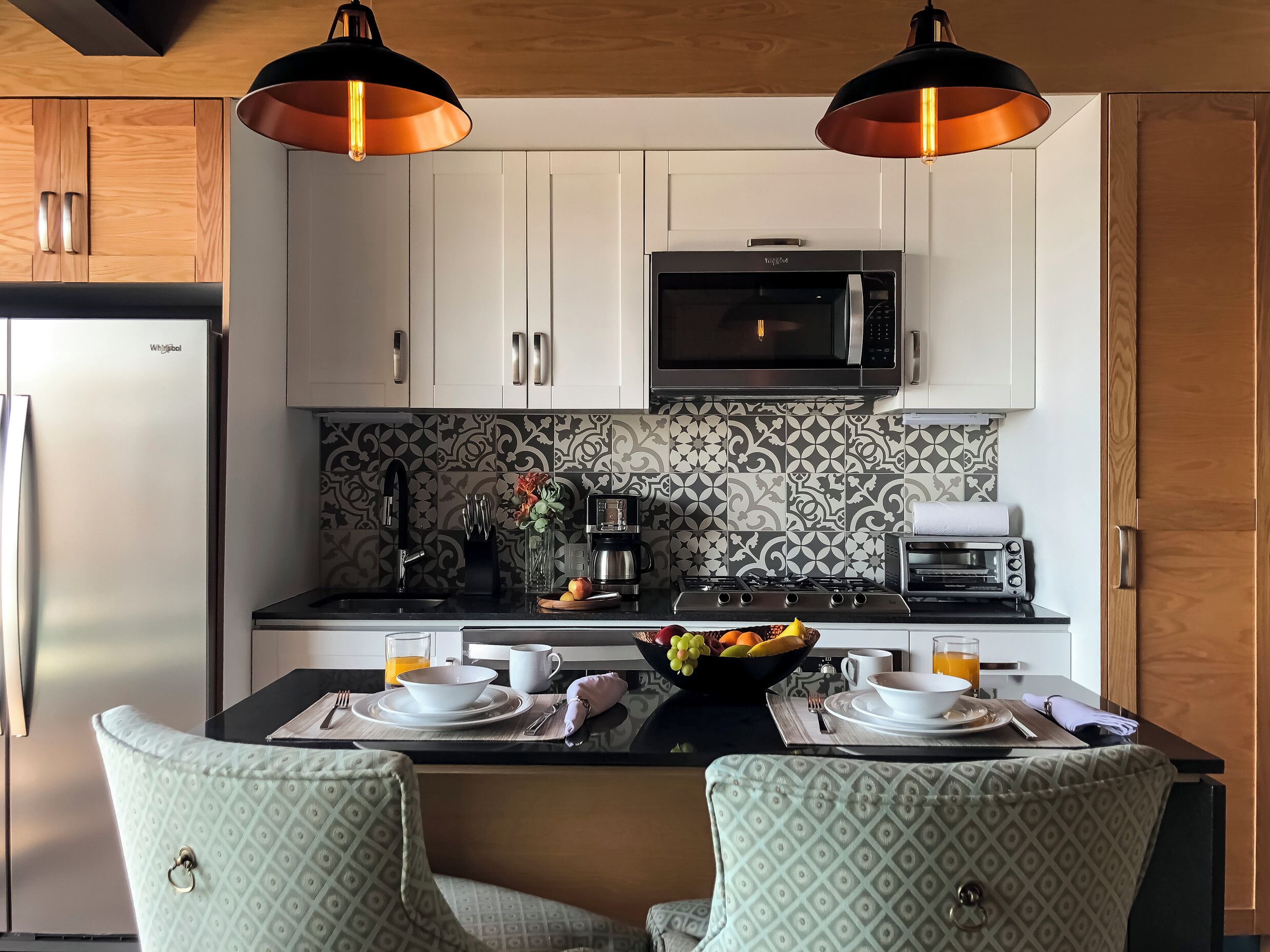 A modern kitchen with white cabinets, a microwave, a coffee maker, and a toaster. There are two chairs with patterned cushions and a fruit bowl on the counter.