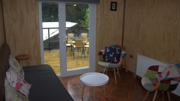 A room with a sofa, a small round table, and two chairs. There is a sliding glass door leading to an outdoor area with a table and chairs. - El Refugio de la Cuenca (Puerto Octay)