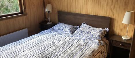 A bedroom with a wooden headboard, a patterned bedspread, two pillows, a bedside table with a lamp, and a window with a view of trees.