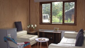 A living room with a sofa, a small table with cookies, a TV, and a window with a view of trees. - El Refugio de la Cuenca (Puerto Octay)