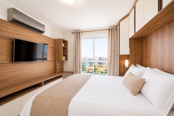 A hotel room with a large bed, a flat-screen TV mounted on a wooden wall, a window with curtains, and a view of the cityscape.