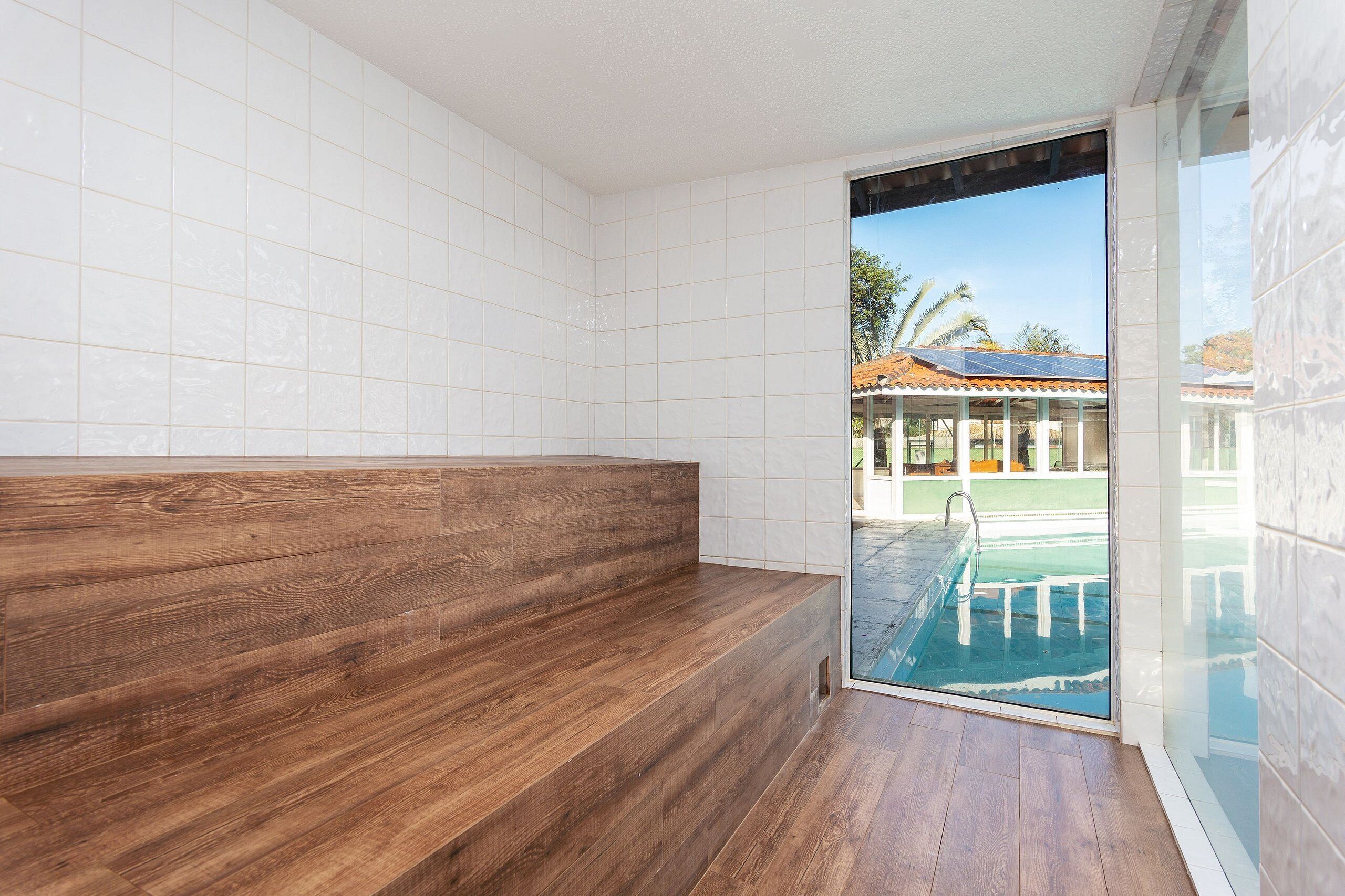 A bathroom with white tiled walls, a wooden bench, and a glass door leading to a pool area.