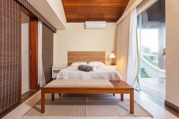 A bedroom with a wooden bed, a bedside table with a lamp, a window with curtains, and an air conditioning unit. - 40m from the beach! Beautiful house with private pool BZ26 (Armação dos Búzios)