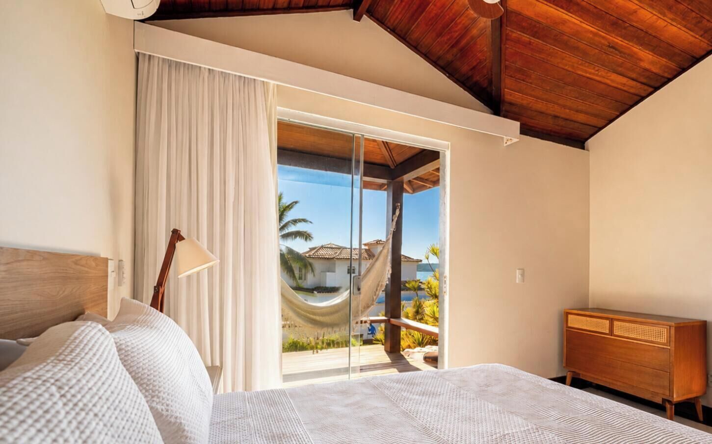 Une chambre avec un lit, une table de chevet en bois, une grande fenêtre avec des rideaux et une vue sur une piscine et des palmiers.