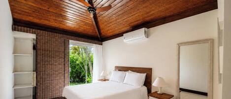 A bedroom with a wooden ceiling, a bed with white linens, a bedside table with a lamp, and a window with a view of greenery.