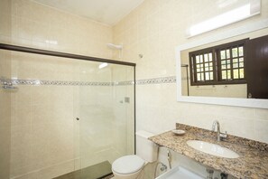 Un baño con ducha envuelta en vidrio, inodoro blanco, lavabo con encimera de granito y espejo con ventana arriba.