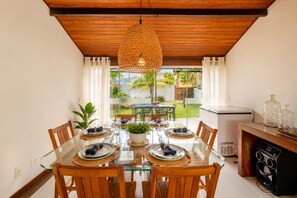 A dining area with a glass table, wooden chairs, a hanging light fixture, and a view of a garden through large windows. - Bz04 30 steps from the beach! Beautiful house with a pool! (Armação dos Búzios)