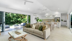 A modern living room with a sofa, coffee table, and dining area. There is a kitchen in the background with a refrigerator and a dining table.