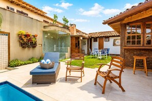 Zona junto a la piscina con una tumbona, una mesa de madera y sillas, ducha con paredes de vidrio y un pequeño jardín con cactus.