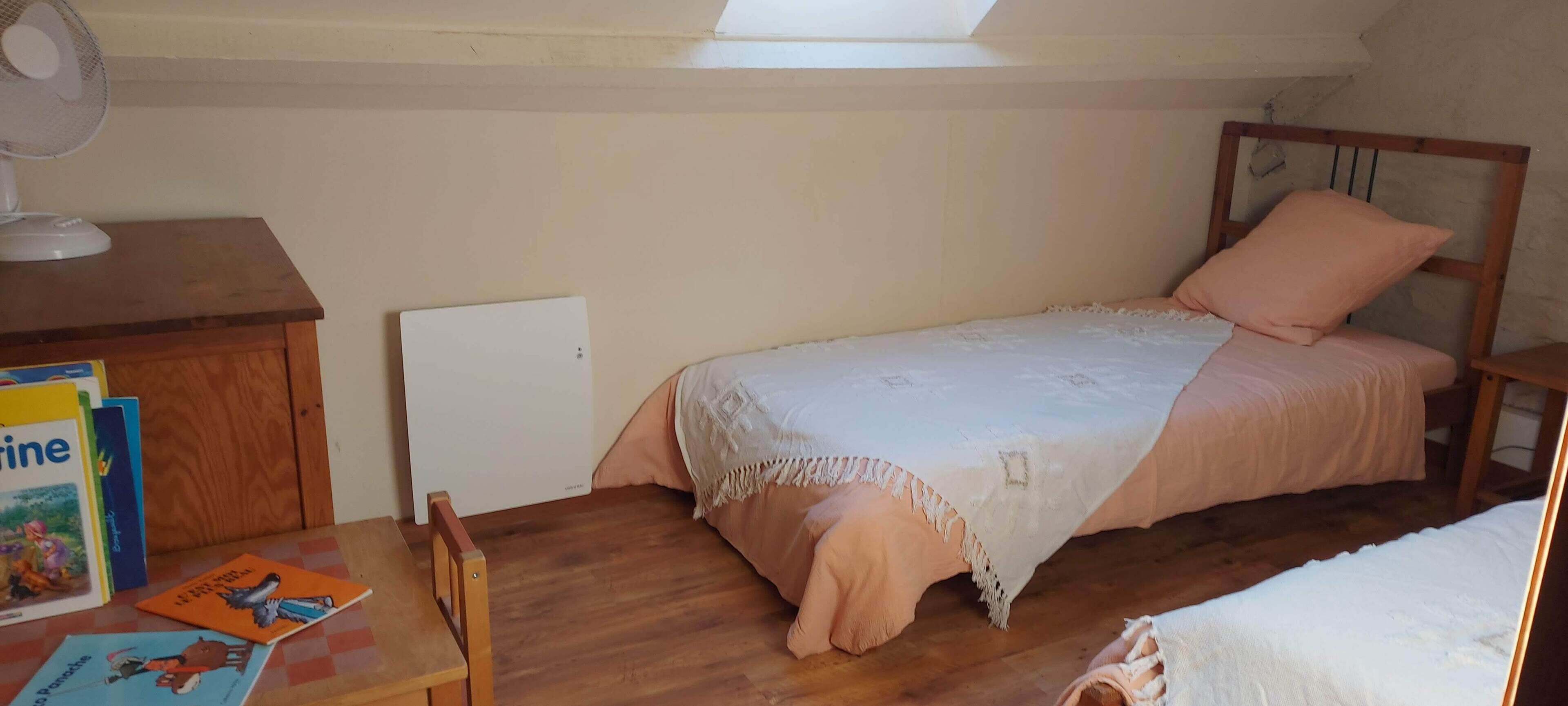 A small, cozy bedroom with a single bed, a wooden nightstand, a desk with books, and a ceiling fan.