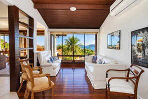 Ein großzügiger Wohnbereich mit einem weißen Sofa, Holzstühlen und einem großen Fenster mit Blick auf Palmen und das Meer.