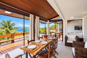 Een eetgedeelte met houten tafels en stoelen, uitzicht op de oceaan en een balkon met houten hekwerk.