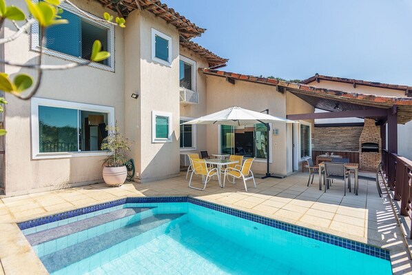 A swimming pool with tiled edges, a patio area with outdoor furniture, and a brick fireplace.
