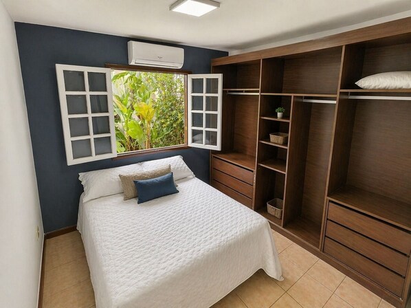 Une chambre à coucher avec un lit, une grande fenêtre dont les volets sont ouverts, une armoire et un climatiseur fixé au plafond.