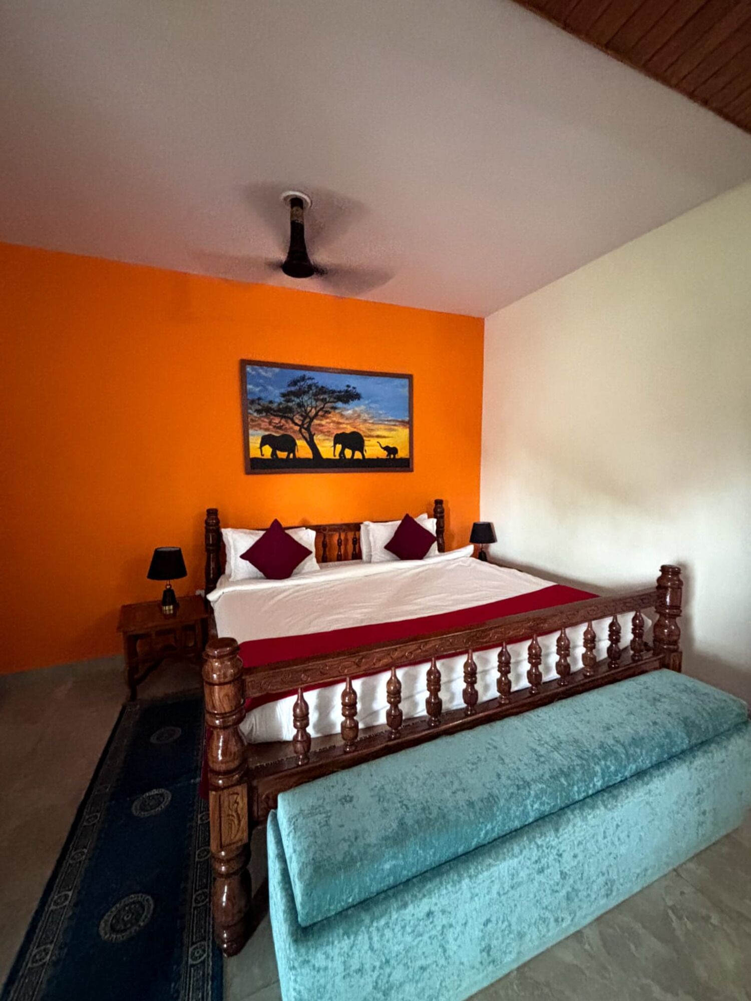 Un dormitorio con pared naranja, una cama de madera con colcha roja y blanca, un banco azul y un cuadro de elefantes.