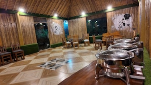Un espace repas doté d’un toit de chaume, de murs en bambou et d’un sol carrelé. On y trouve plusieurs chaises et tables en bois, ainsi qu’un buffet avec des plats de service en métal.