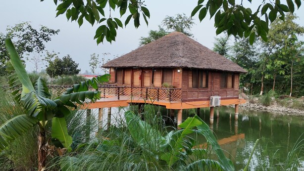 Une maison en bois avec un toit de chaume, construite sur pilotis au-dessus de l’eau, et entourée de verdure.