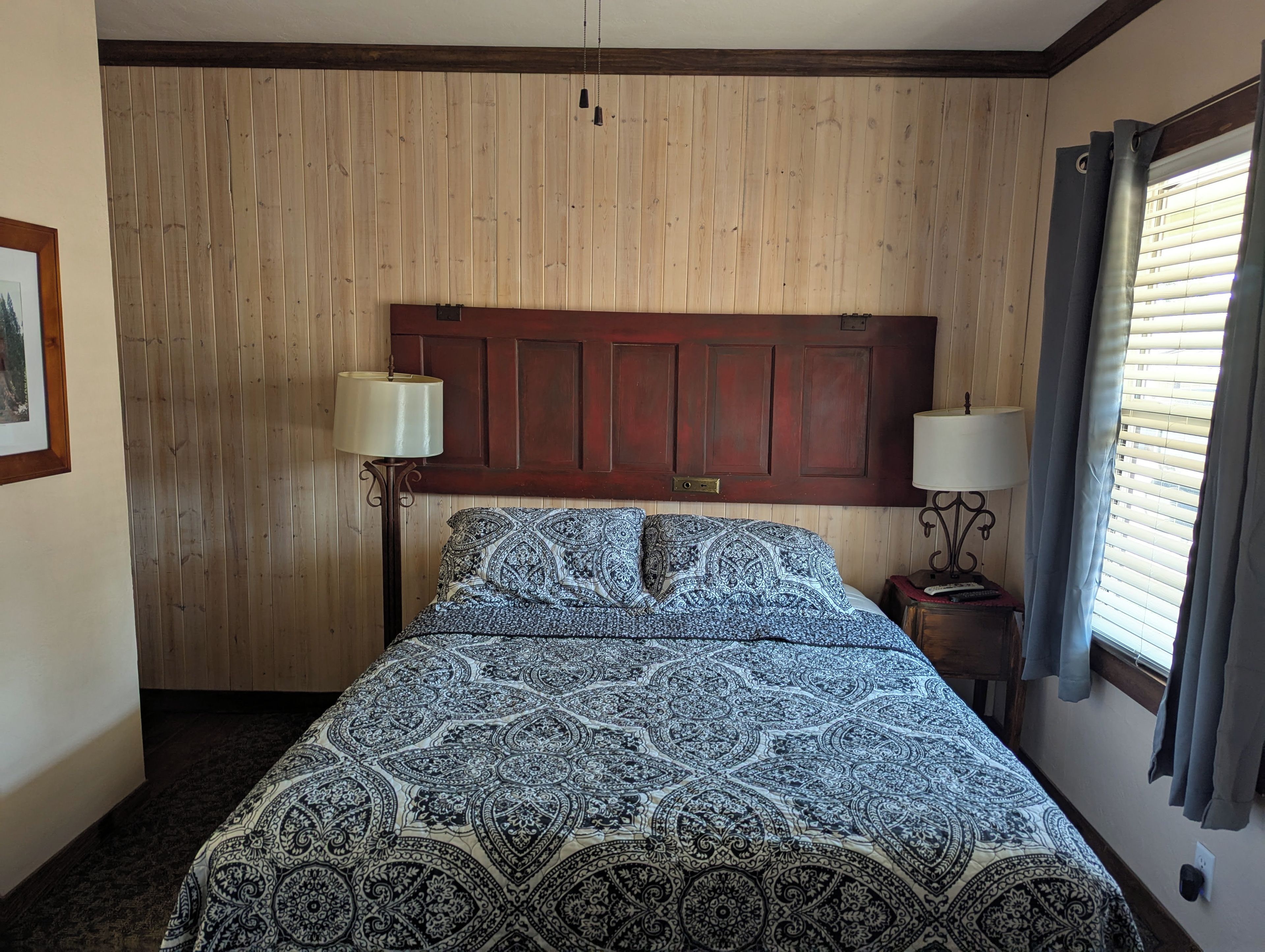 Un dormitorio con cabecera de madera, colcha estampada, dos lámparas de noche y una ventana con persianas.